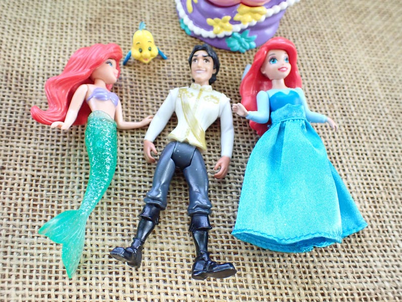 May include: Ariel and Prince Eric dolls from the Disney film, The Little Mermaid. Ariel has a green tail and red hair, and Prince Eric wears a white shirt and black trousers. A small yellow fish is also present.