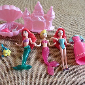 May include: A collection of pink plastic toys featuring mermaid dolls, a castle, a seashell with jewelry, and a yellow and blue fish figurine. The mermaid dolls have red, blonde, and pink hair and wear colorful tops and tails.