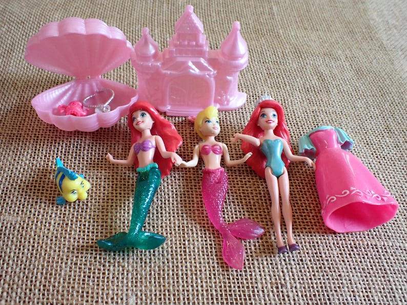 May include: A collection of pink plastic toys featuring mermaid dolls, a castle, a seashell with jewelry, and a yellow and blue fish figurine. The mermaid dolls have red, blonde, and pink hair and wear colorful tops and tails.
