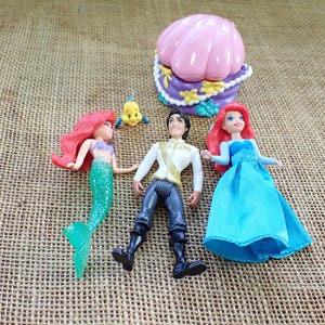 May include: A selection of Disney's The Little Mermaid figurines, featuring Ariel, Prince Eric, and Flounder. Ariel has a green tail and red hair, Prince Eric wears a white jacket, and Flounder is yellow. A pink seashell-shaped container is also present.