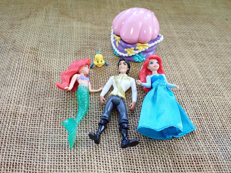 May include: A selection of Disney's The Little Mermaid figurines, featuring Ariel, Prince Eric, and Flounder. Ariel has a green tail and red hair, Prince Eric wears a white jacket, and Flounder is yellow. A pink seashell-shaped container is also present.