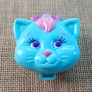 May include: A light blue cat-shaped toy with pink accents. The toy features large blue eyes, long eyelashes, and a pink nose. A pink bow and a small circular emblem are on top of the cat's head. The toy is on a textured surface.