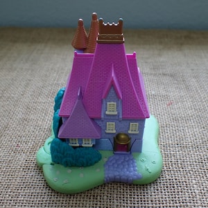 May include: A plastic toy house featuring a purple roof with brick-like textures, light purple walls, and a brown turret. The structure sits on a light green base with molded teal bushes and a purple path leading to a small, arched doorway.