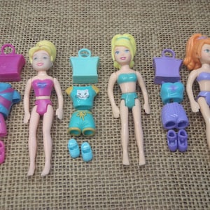 Op de afbeelding: Vier miniatuurpoppen met verwisselbare outfits en accessoires. Elke pop heeft een ander kapsel en draagt een badpak. De outfits omvatten tops, shorts, schoenen en handtassen in verschillende kleuren zoals roze, blauw en paars.