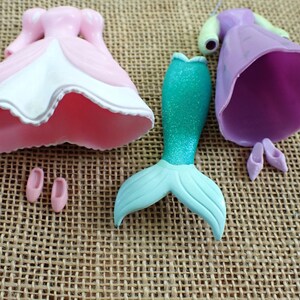 May include: A collection of doll parts, including a pink and white dress, a mermaid tail with a teal glitter finish, a purple dress, and two pairs of small shoes. The items are arranged on a textured, tan surface.