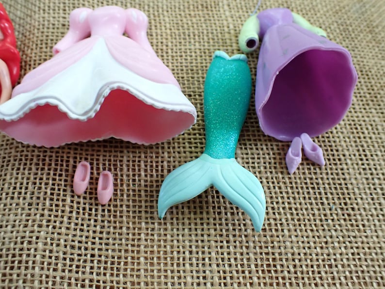 May include: A collection of doll parts, including a pink and white dress, a mermaid tail with a teal glitter finish, a purple dress, and two pairs of small shoes. The items are arranged on a textured, tan surface.