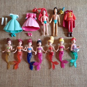 May include: A collection of miniature dolls and accessories, including mermaid figures with colorful tails, dresses, and a prince figure. The dolls have various hair colors and are arranged on a textured surface.