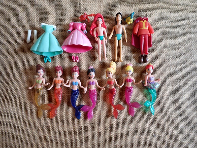 May include: A collection of miniature dolls and accessories, including mermaid figures with colorful tails, dresses, and a prince figure. The dolls have various hair colors and are arranged on a textured surface.
