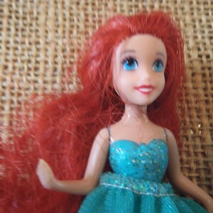 May include: A small doll with bright red hair and blue eyes. The doll is wearing a teal dress with glitter accents. The doll's skin is a light tone, and it has a smiling expression. The background is a woven, tan material.