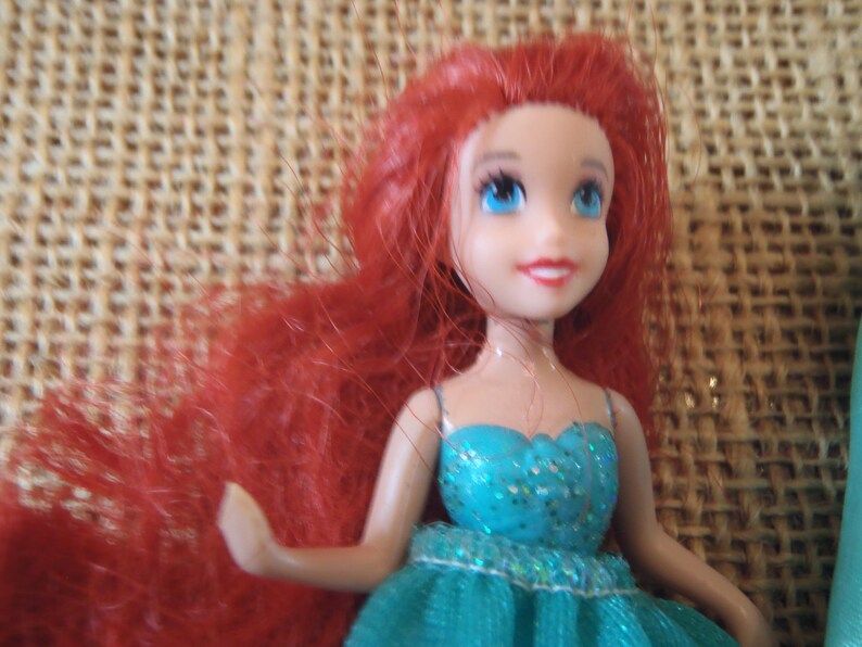 May include: A small doll with bright red hair and blue eyes. The doll is wearing a teal dress with glitter accents. The doll's skin is a light tone, and it has a smiling expression. The background is a woven, tan material.