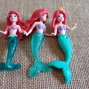 Puede incluir: Tres figuras de sirenas con cabello rojo, tops de conchas moradas y colas turquesas. Una sirena lleva una corona dorada. Las mu&ntilde;ecas est&aacute;n posadas sobre una superficie texturizada y beige.
