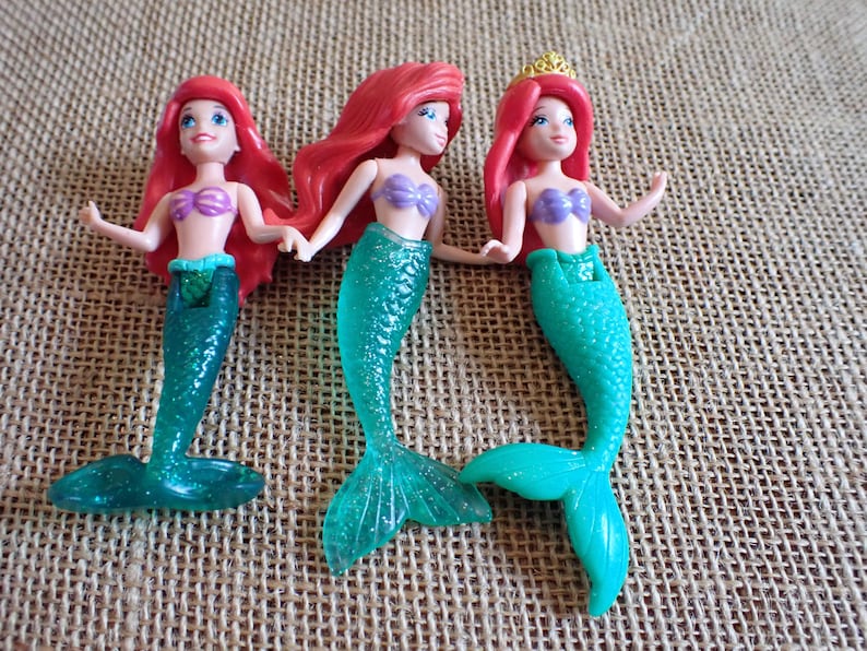Puede incluir: Tres figuras de sirenas con cabello rojo, tops de conchas moradas y colas turquesas. Una sirena lleva una corona dorada. Las mu&ntilde;ecas est&aacute;n posadas sobre una superficie texturizada y beige.