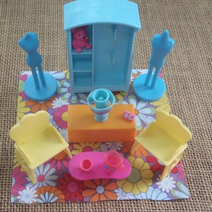 May include: A blue toy dollhouse with a pink teddy bear inside, two blue doll stands, two yellow chairs, a pink table with two cups, and an orange table with a blue vase on a colourful floral rug.