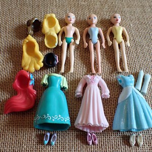 May include: A collection of miniature doll figures and accessories. The dolls are nude with painted features. Accessories include dresses in teal, pink, and blue, along with shoes and wigs in various colors. The items are arranged on a textured surface.