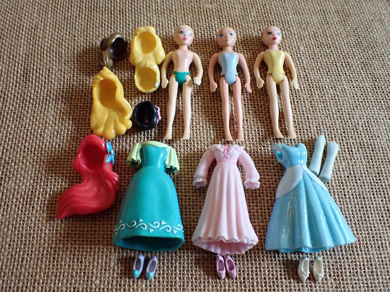 May include: A collection of miniature doll figures and accessories. The dolls are nude with painted features. Accessories include dresses in teal, pink, and blue, along with shoes and wigs in various colors. The items are arranged on a textured surface.