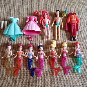 May include: A collection of small plastic dolls and accessories. The set includes mermaid dolls with colorful tails, male and female dolls, dresses, and other clothing items. The dolls have various hair colors and skin tones. The accessories include shoes and a red cape.