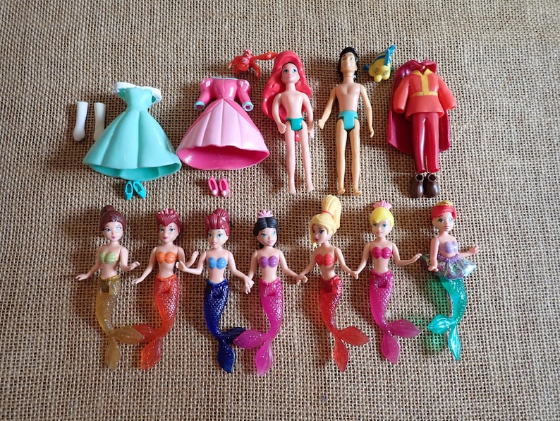 May include: A collection of small plastic dolls and accessories. The set includes mermaid dolls with colorful tails, male and female dolls, dresses, and other clothing items. The dolls have various hair colors and skin tones. The accessories include shoes and a red cape.