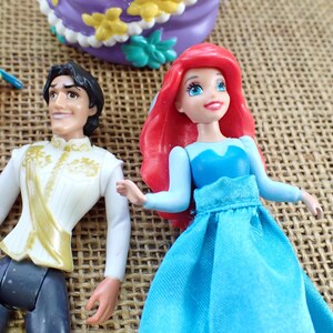 May include: Ariel and Prince Eric figurines with a yellow fish and a purple cake. Ariel has red hair and a blue dress. Prince Eric wears a white jacket with gold accents. The cake is purple with yellow and green decorations.