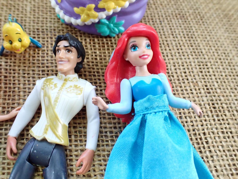 May include: Ariel and Prince Eric figurines with a yellow fish and a purple cake. Ariel has red hair and a blue dress. Prince Eric wears a white jacket with gold accents. The cake is purple with yellow and green decorations.