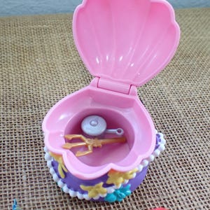 May include: A pink clamshell toy, open to show a gold trident and a silver disc. The shell is trimmed with purple, white, and yellow details. The toy is designed to open and close, resembling a treasure chest.