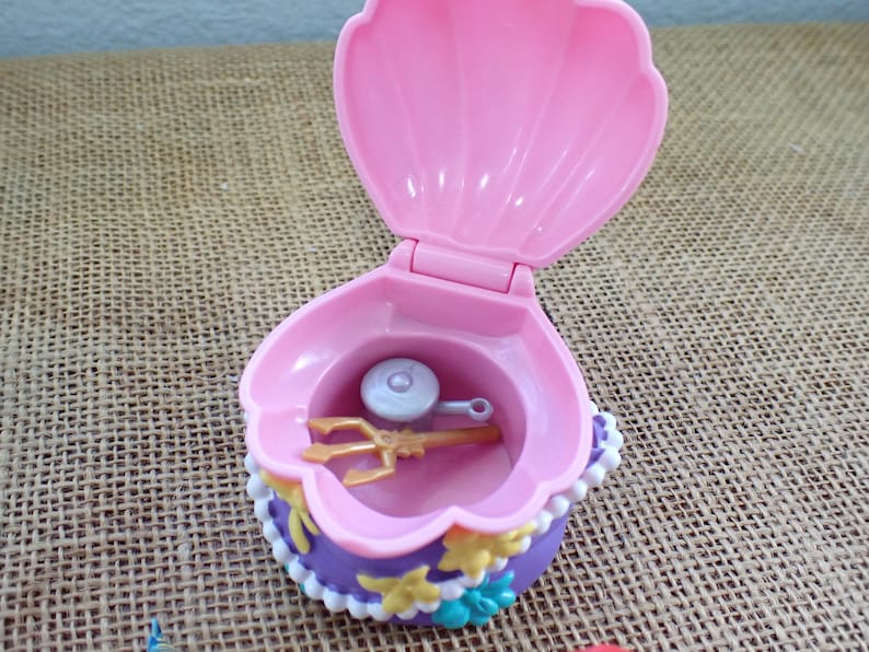 May include: A pink clamshell toy, open to show a gold trident and a silver disc. The shell is trimmed with purple, white, and yellow details. The toy is designed to open and close, resembling a treasure chest.