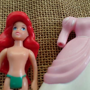 May include: Ariel doll with red hair, blue eyes, and a green bikini bottom. Beside her is a pink and white princess dress. The doll is made of plastic and is a toy.