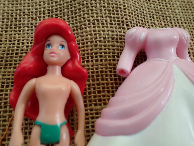 May include: Ariel doll with red hair, blue eyes, and a green bikini bottom. Beside her is a pink and white princess dress. The doll is made of plastic and is a toy.