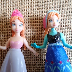 Puede incluir: Dos peque&ntilde;as mu&ntilde;ecas con tem&aacute;tica de princesa. Una mu&ntilde;eca viste un vestido rosa y blanco con una corona rosa. La otra mu&ntilde;eca lleva un vestido azul con un top negro y un tocado rosa y azul. Ambas mu&ntilde;ecas tienen pelo pelirrojo y ojos azules.