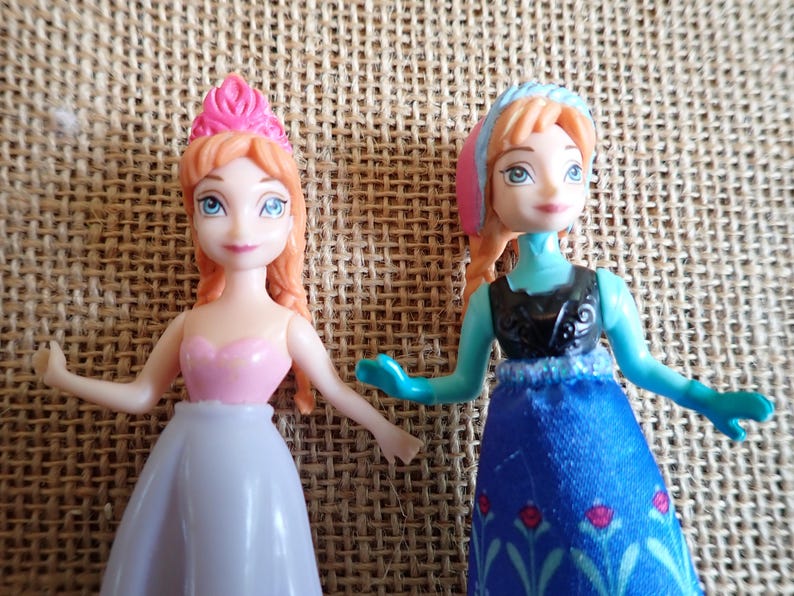 Puede incluir: Dos peque&ntilde;as mu&ntilde;ecas con tem&aacute;tica de princesa. Una mu&ntilde;eca viste un vestido rosa y blanco con una corona rosa. La otra mu&ntilde;eca lleva un vestido azul con un top negro y un tocado rosa y azul. Ambas mu&ntilde;ecas tienen pelo pelirrojo y ojos azules.