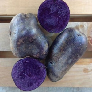May include: Four purple potatoes, two whole and two cut in half, showing the vibrant purple flesh. The potatoes are arranged on a wooden surface.