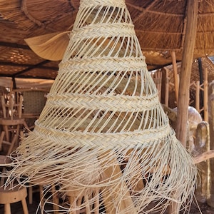 May include: A woven lampshade made of natural materials hangs from a thatched roof. The lampshade is cone-shaped with a fringe of woven strands. The lampshade is hanging in a room with wooden furniture.