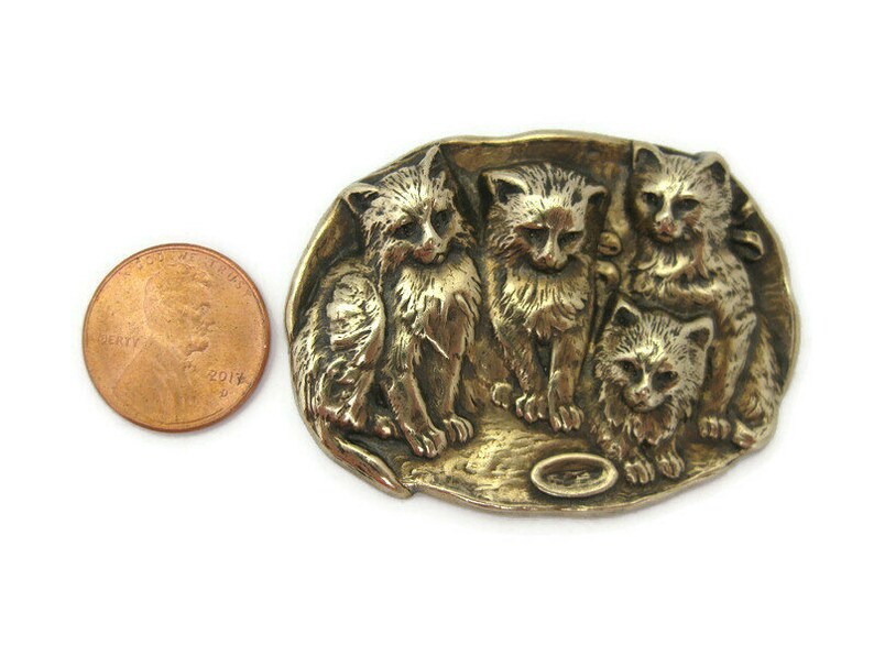 Peut inclure: Une broche en m&eacute;tal dor&eacute; repr&eacute;sentant quatre chatons dans une sc&egrave;ne ludique. Les chatons sont repr&eacute;sent&eacute;s en relief, en trois dimensions.