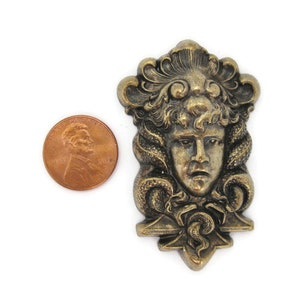 May include: A gold-toned metal pendant featuring a detailed sculpted face with a crown of leaves and snakes. The pendant is about the size of a US penny.