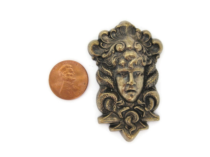 May include: A gold-toned metal pendant featuring a detailed sculpted face with a crown of leaves and snakes. The pendant is about the size of a US penny.