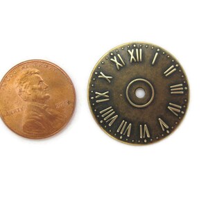 May include: A close-up of a brass clock face with Roman numerals. The clock face is about the size of a US penny.
