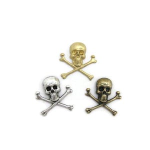 May include: Three skull and crossbones charms in gold, silver, and bronze tones. Each skull has detailed facial features and is positioned above two crossed bones. The charms are set against a white background.