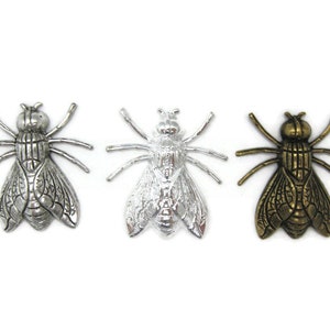 Op de afbeelding: Drie metalen vlieg charms in zilver, wit en goud. Elke charm heeft een gedetailleerd ontwerp van een vlieg met ingewikkelde vleugels en poten.