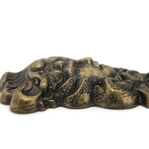May include: A gold-toned metal wall decor featuring a detailed, sculpted face with intricate hair and a textured, scaly design.