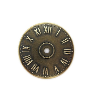 May include: A round, antique gold-toned clock face with Roman numeral markings. The clock face has a small hole in the center.