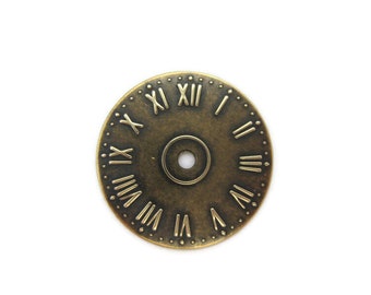 May include: A round, antique gold-toned clock face with Roman numeral markings. The clock face has a small hole in the centre.