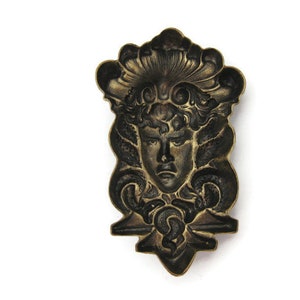 May include: A vintage brass decorative plaque featuring a detailed relief of a woman's face with a crown of leaves and flowers. The plaque has a dark patina and intricate details.