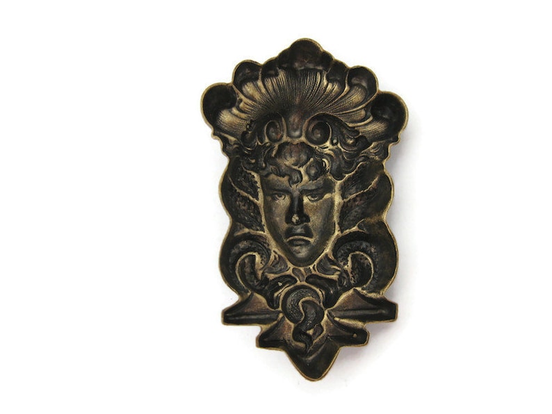 May include: A vintage brass decorative plaque featuring a detailed relief of a woman's face with a crown of leaves and flowers. The plaque has a dark patina and intricate details.