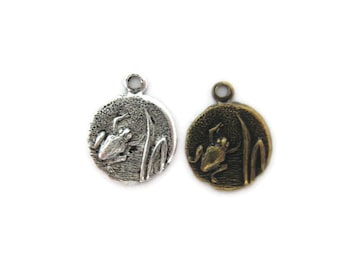 May include: Two silver and bronze frog charms with a detailed frog design on a round pendant. The frog is sitting on a lily pad with reeds.