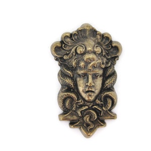 May include: A gold-toned metal brooch featuring a detailed sculpted face with a crown of leaves and snakes. The face has a serious expression with a furrowed brow and a slightly open mouth.