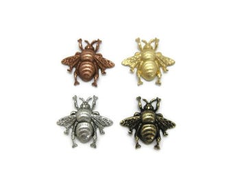 May include: Four metal bee-shaped embellishments in bronze, gold, silver, and antique brass. Each bee has detailed wings and body segments, perfect for crafting or jewelry making. The bees are arranged on a white background.