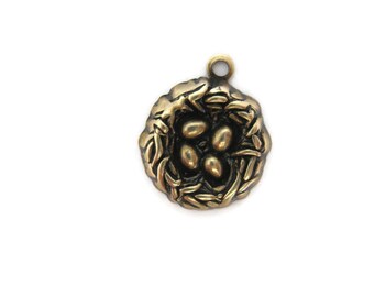May include: A bronze-colored charm featuring a detailed bird's nest design. The nest is filled with four oval-shaped eggs. The charm has a loop at the top for attaching to a necklace or bracelet. The overall design is detailed and textured.