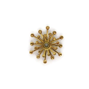 May include: A gold-toned metal brooch in the shape of a starburst with a central circle. The brooch has a geometric design with multiple points radiating outwards.