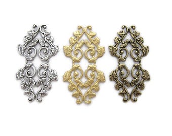 Op de afbeelding: Drie sierlijke metalen filigraanversieringen in zilver, goud en brons. Elk stuk heeft een delicaat bloemmotief met ingewikkelde spiralen en bladeren.