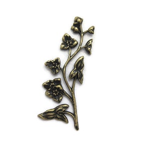 May include: A bronze-colored metal floral embellishment featuring a detailed design of flowers and leaves. The decorative piece has a vintage aesthetic and is likely used for crafting or jewelry making. The floral element is set against a white background.