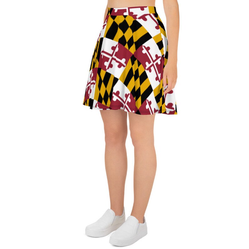 May include: A skater skirt featuring a geometric pattern in black, gold, white, and burgundy. The design incorporates a repeating pattern of diamonds and crosses. The skirt is worn with white slip-on shoes.